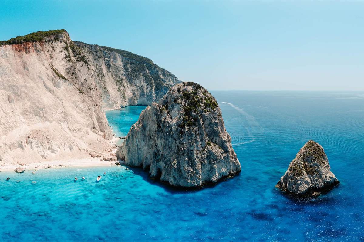Zakynthos Mizithres and Eros Cave boat tour: a tall grey rock topped with green scrub rising from deep blue sea beside a smaller rock, with a pale limestone cliff and a thin cream beach behind them under a clear sky
