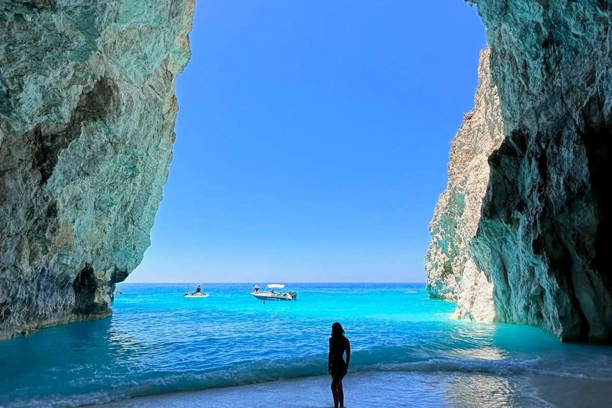 Zakynthos Mizithres and Eros Cave boat tour: a tall opening between two rough grey-green rock walls framing bright turquoise sea and a clear blue sky, with the dark outline of one person on the sandy shore in front and two small boats far out on the water