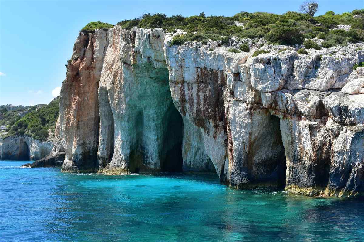 Zakynthos shipwreck beach and blue caves tour: pale limestone sea caves and rock arches of the Blue Caves at Cape Skinari rising from clear turquoise water, topped with green scrub under a blue sky