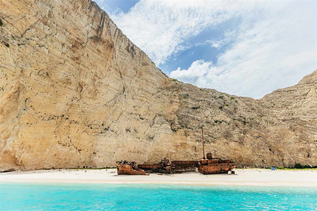 Zakynthos shipwreck beach and blue caves tour: the rusted wreck lying on the white sand of Navagio beach below a tall pale limestone cliff, with bright turquoise water in the foreground and a blue sky with soft clouds