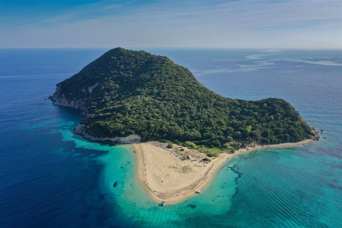 Zakynthos turtles Marathonisi and Keri Caves boat tour: Marathonisi island seen from above, a green wooded hill with pale rock at its edge and a curved cream sand spit reaching into clear turquoise water, with deep blue sea and a soft sky behind