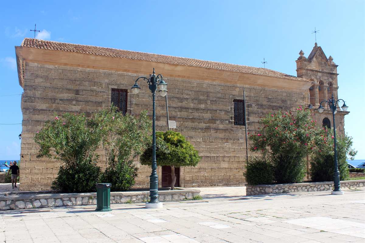 Zante tours: the sandstone Church of Saint Nikolaos on Solomos Square in Zakynthos Town, with its bell tower at the right, iron street lamps and pink oleander bushes in front, and a strip of blue sea far left under a clear sky